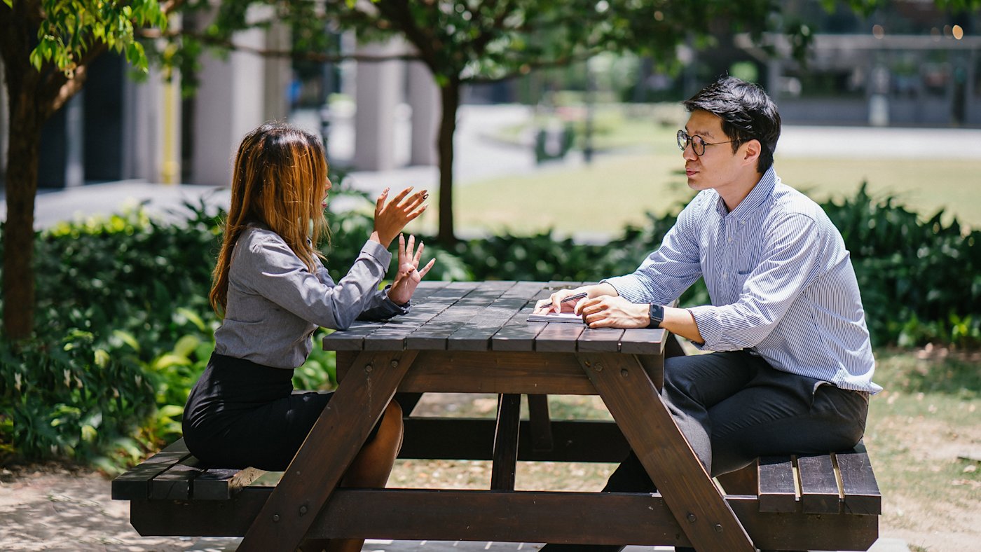 Two professionals in business-casual clothing talking at a wooden picnic table under leafy trees